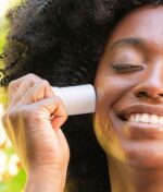 En person med krøllet hår smiler med lukkede øjne, mens de påfører COOLA Refreshing Water Stick SPF 50 - 22 g på kinden.