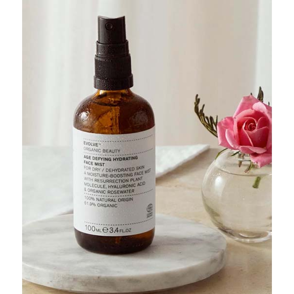 En brun flaske Evolve Age Defying Hydrating Face Mist 100 ml placeres på en marmorbakke med en lyserød rose i en klar vase ved siden af.