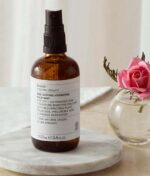 En brun flaske Evolve Age Defying Hydrating Face Mist 100 ml placeres på en marmorbakke med en lyserød rose i en klar vase ved siden af.