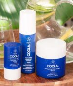 Tre COOLA hudplejeprodukter med blå beholdere: COOLA Refreshing Water Stick SPF 50 - 22 g, Refreshing Water Mist og Refreshing Water Cream, vises på et bord ved siden af en klar glaskande med citronskiver.