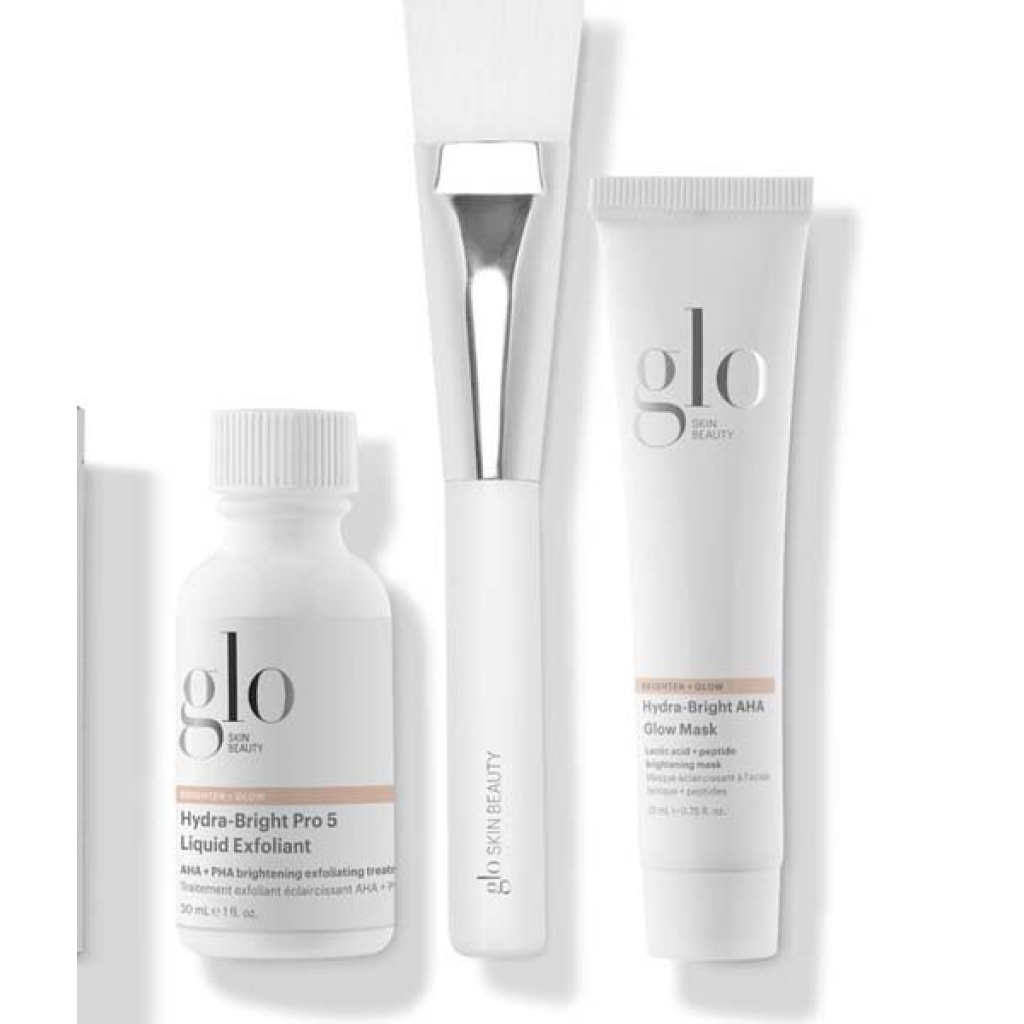 Three Glo Skin Beauty Radiant Spa Day Kit - Begrænsede hudplejeprodukter: en hvid flaske Hydra-Bright Pro 5 Liquid Exfoliant, en hvid tube Hydra-Bright AHA Glow Mask og en hvid påføringsbørste.
