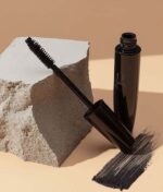 Open Glo Beauty Time To Shine - Warm Collection, placerer mascaratuben med børste ved siden af en tekstureret sten og et mascarastrøg på en beige overflade.