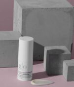 En hvid cylindrisk flaske Glo Skin Beauty Revitalize Eye Care Duo 2 x 15 ml placeres på en lyserød overflade med grå betonklodser i baggrunden; en klat af serummet er ved siden af flasken.