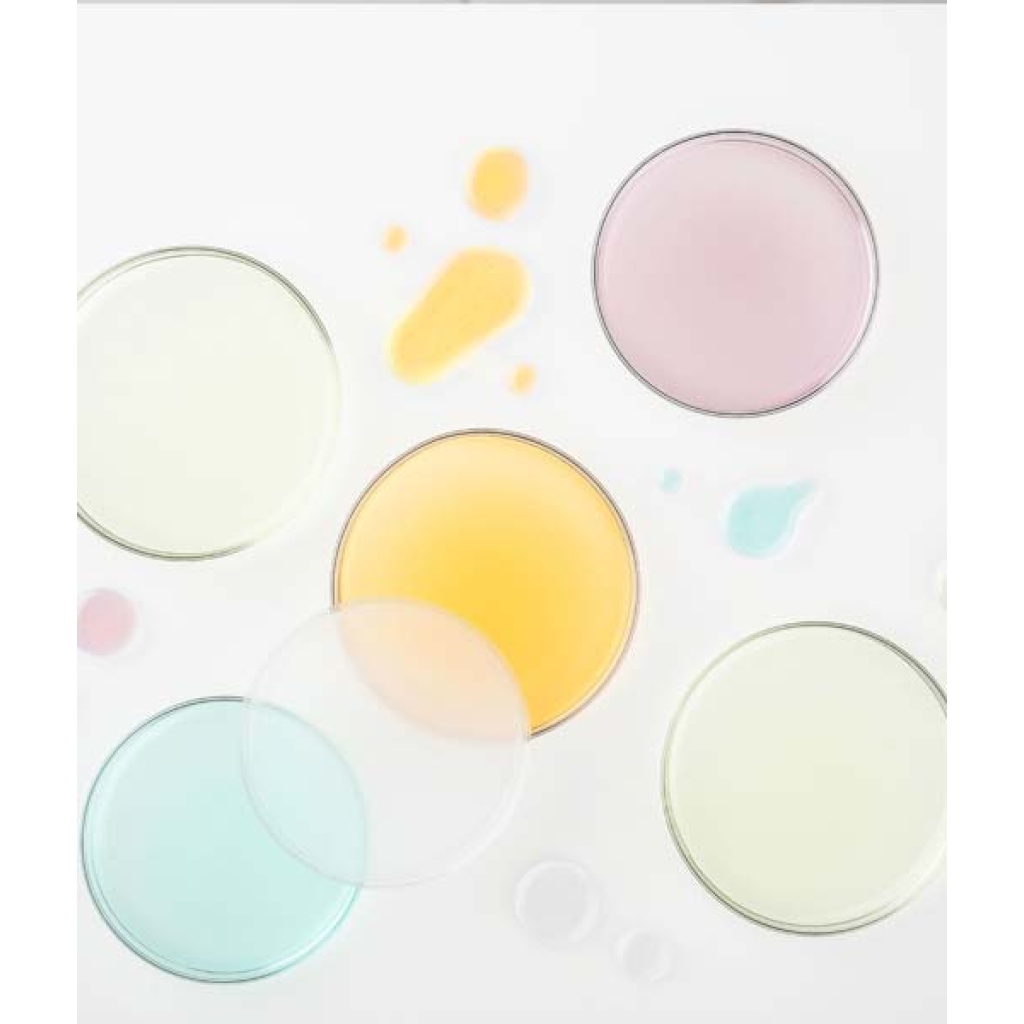 Fem petriskåle indeholdende Glo Hydra-Bright Pro 5 Liquid Exfoliant i lyserøde, gule, aqua og klare varianter er arrangeret på en hvid overflade. Dråber af samme farvede eksfolierende væsker er spredt rundt om dem.