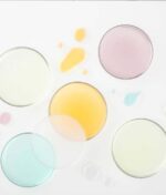 Fem petriskåle indeholdende Glo Hydra-Bright Pro 5 Liquid Exfoliant i lyserøde, gule, aqua og klare varianter er arrangeret på en hvid overflade. Dråber af samme farvede eksfolierende væsker er spredt rundt om dem.