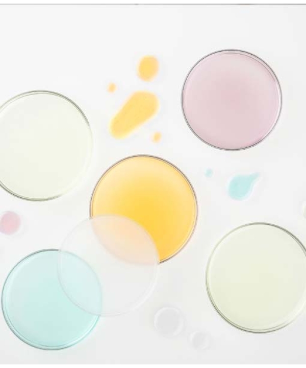 Fem petriskåle indeholdende Glo Hydra-Bright Pro 5 Liquid Exfoliant i lyserøde, gule, aqua og klare varianter er arrangeret på en hvid overflade. Dråber af samme farvede eksfolierende væsker er spredt rundt om dem.