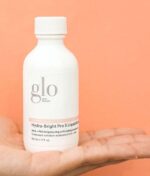 En hvid flaske Glo Hydra-Bright Pro 5 Liquid Exfoliant 60 ml holdes i hånden mod en orange baggrund.