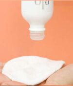 En hånd holder en rund vatrondell, mens Glo Hydra-Bright Pro 5 Liquid Exfoliant i sin hvide flaske dispenserer væske på puden mod en orange baggrund.