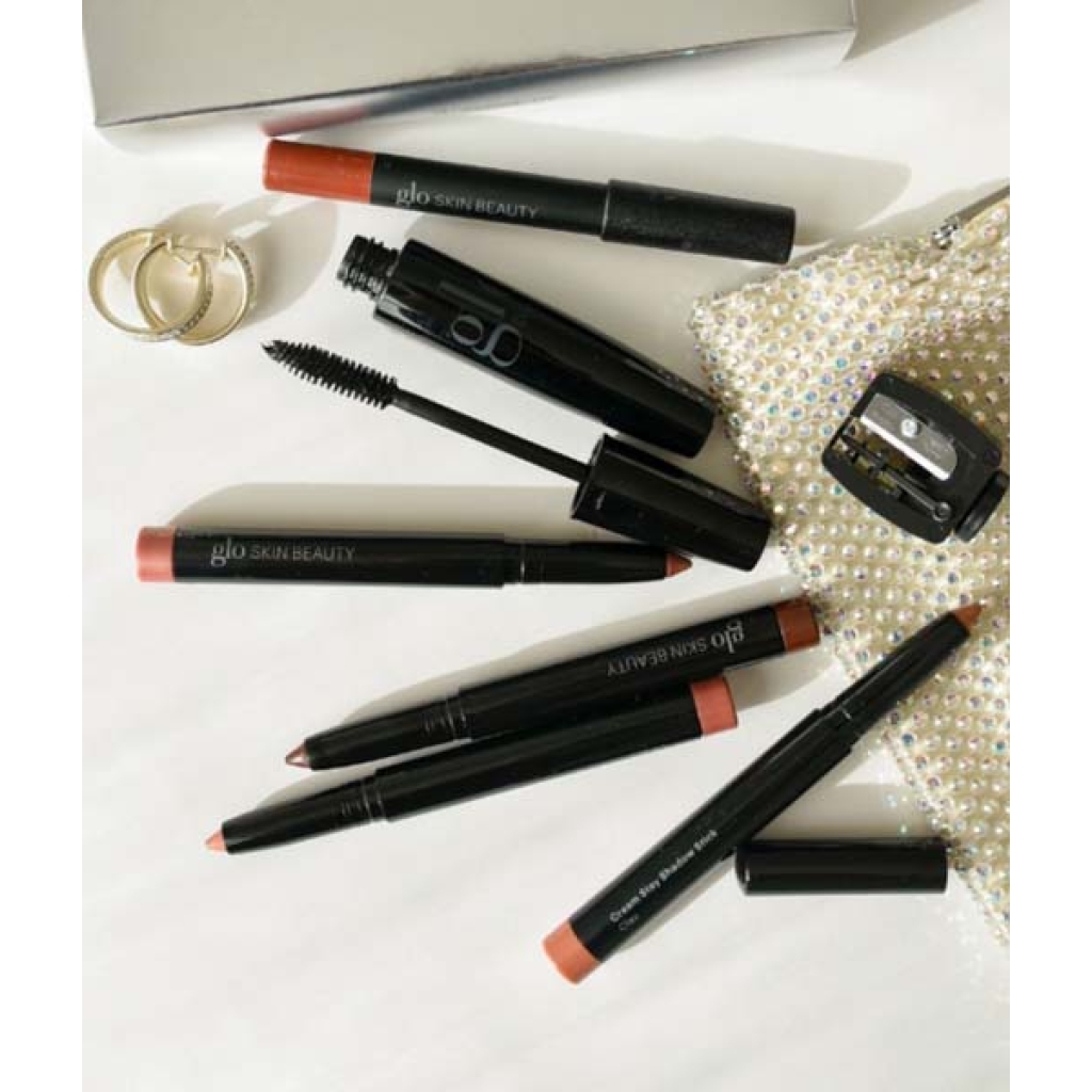 Makeup-artikler fra Glo Beauty Time To Shine - Warm Collection, som omfatter læbeblyanter, en mascara-stav og en blyantspidser, vises på en hvid overflade sammen med to ringe og en lille, glitrende taske.