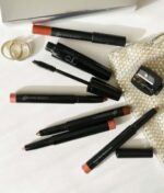 Makeup-artikler fra Glo Beauty Time To Shine - Warm Collection, som omfatter læbeblyanter, en mascara-stav og en blyantspidser, vises på en hvid overflade sammen med to ringe og en lille, glitrende taske.