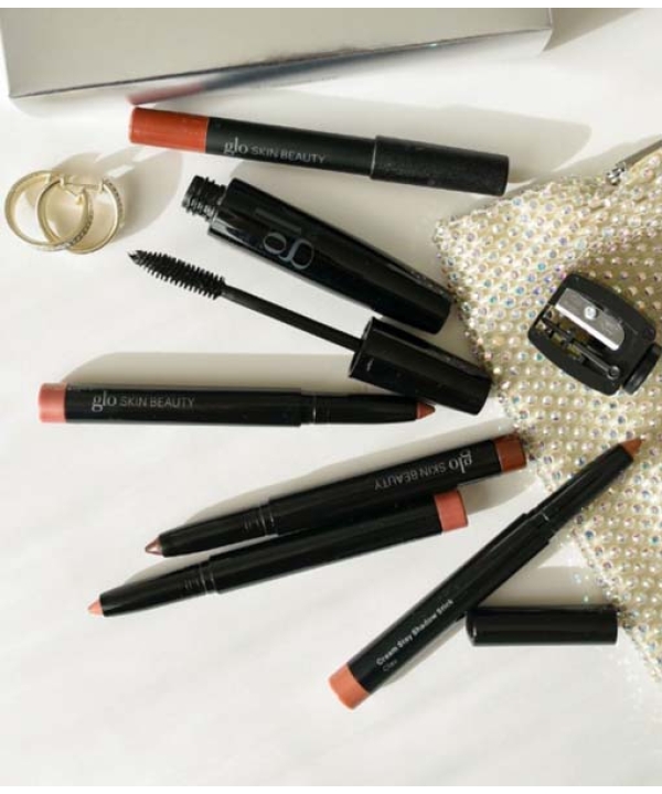 Makeup-artikler fra Glo Beauty Time To Shine - Warm Collection, som omfatter læbeblyanter, en mascara-stav og en blyantspidser, vises på en hvid overflade sammen med to ringe og en lille, glitrende taske.